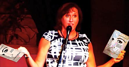 video still of Janet Kuypers holding her books “(pheromemes) 2015-2017 poems” and  “(pheromemes) 2015-2017 show poems” live